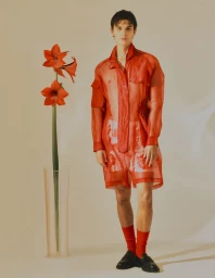 Man wearing a sheer red tunic and matching socks standing beside a tall glass vase with red flowers on a neutral background.