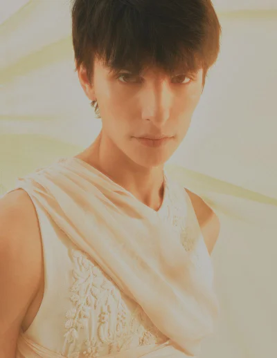 Close-up portrait of a person with short dark hair wearing a white textured top and draped fabric against a warm neutral backdrop