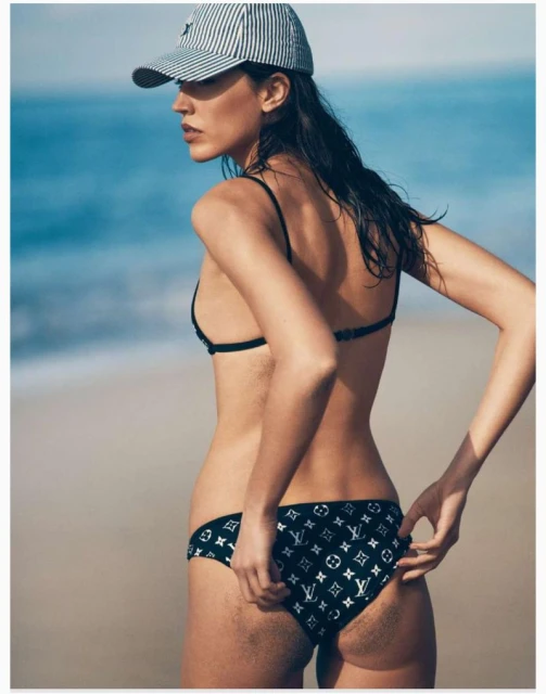 Woman in patterned bikini and striped cap stands on a beach with blue ocean. Summer fashion editorial image with neutral color grading.
