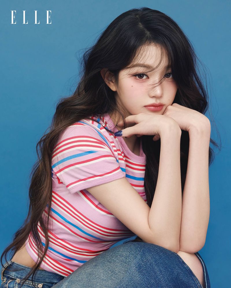 Portrait of a young Asian woman with long wavy dark hair in striped pink top and jeans against a blue backdrop posing for a fashion shot.