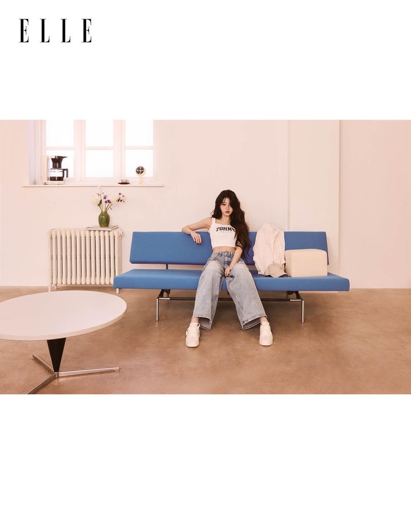 A young woman sits on a blue mid-century modern couch in an indoor setting, wearing a white top and denim pants. Elle logo at top left.