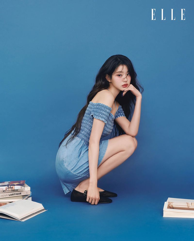 A young adult woman with long dark hair poses in a blue off-the-shoulder dress against a blue background with books and a magazine logo.