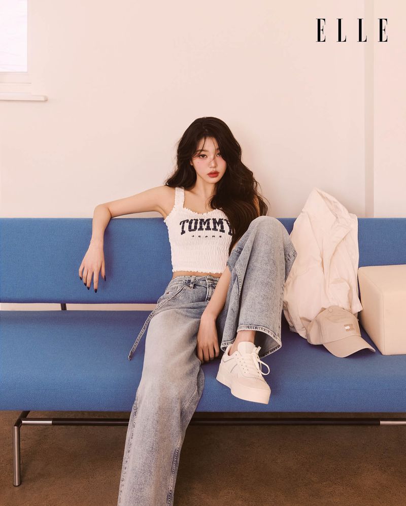 A young woman poses on a blue couch in a studio, wearing a Tommy Hilfiger top and baggy jeans, complemented by white sneakers and a light jacket.
