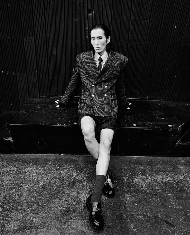 Monochrome portrait of Asian male model in patterned suit, posing outdoors against dark wooden paneling in shiny black loafers.