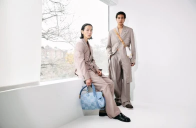 Two male models in beige suits posing indoors near a large window, one holding a blue patterned handbag
