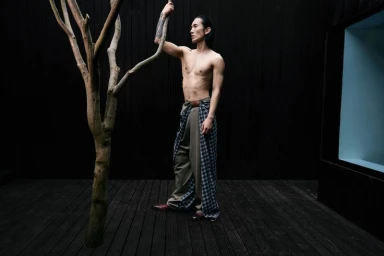 Shirtless male model with long dark hair standing outdoors on dark wood decking next to a bare tree branch, wearing patterned trousers.