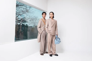 Two male models in beige tailored suits posing in a bright studio with a large window showing a street view.