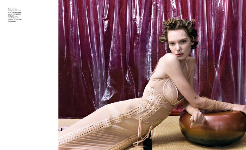 A fashion editorial shot showcases a young woman in a beige corset dress, resting on a brown pouf against a magenta plastic sheet backdrop.