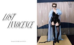 Model wearing black corset and blue feathered cape standing before plastic sheeting for Harper's Bazaar Italia