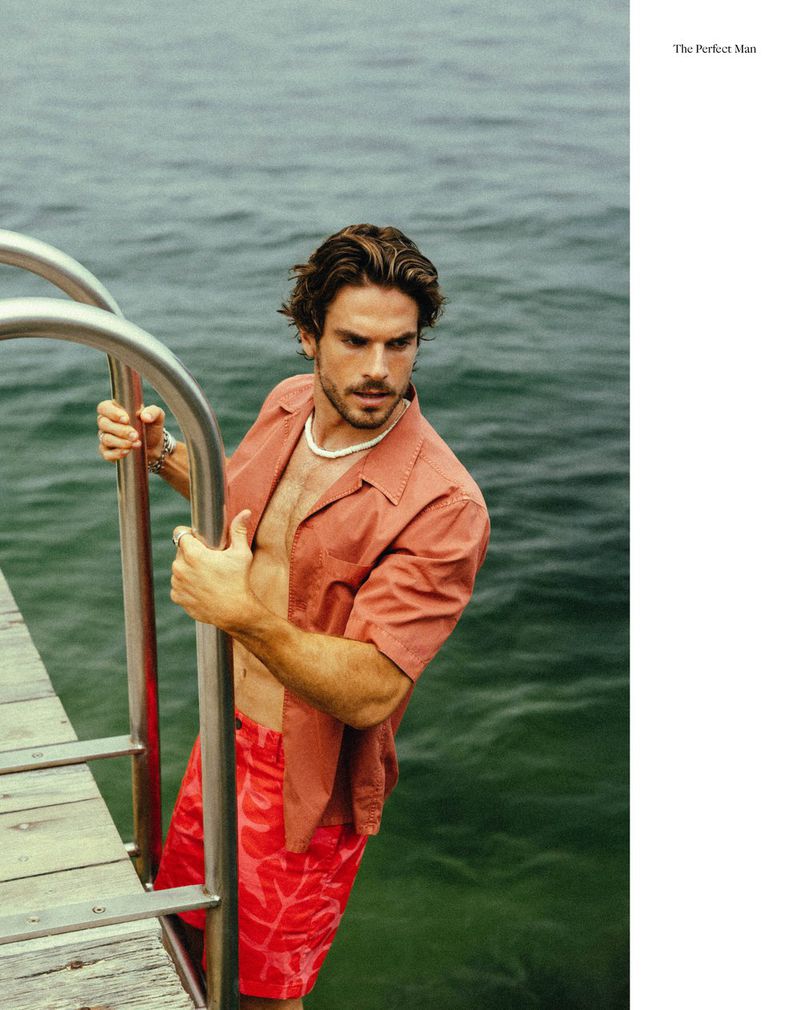 A brunette adult male on a pier wearing coral shirt and red shorts, clutching a silver metal ladder with an ocean background.
