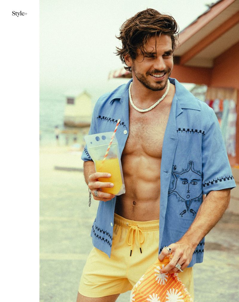A man with a drink wearing an unbuttoned blue shirt and yellow shorts, stands on a beach with a building in the background.