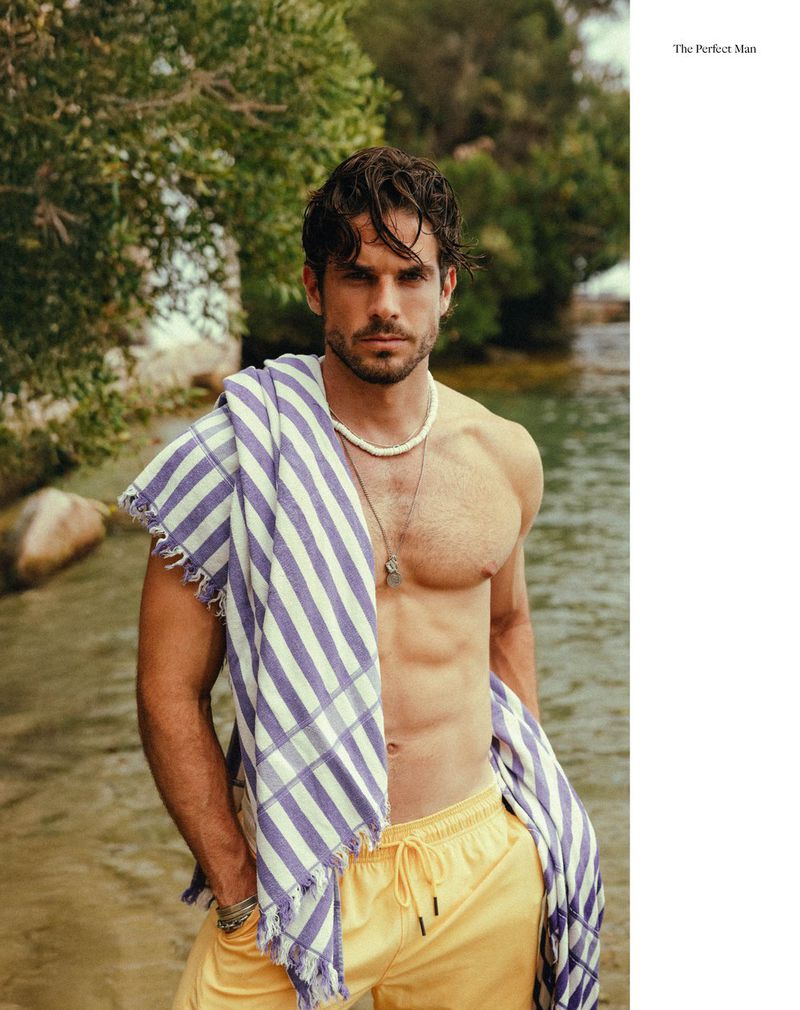 A handsome man with a towel over his shoulder stands outdoors near the water, wearing yellow shorts and necklaces in a portrait-style shot.