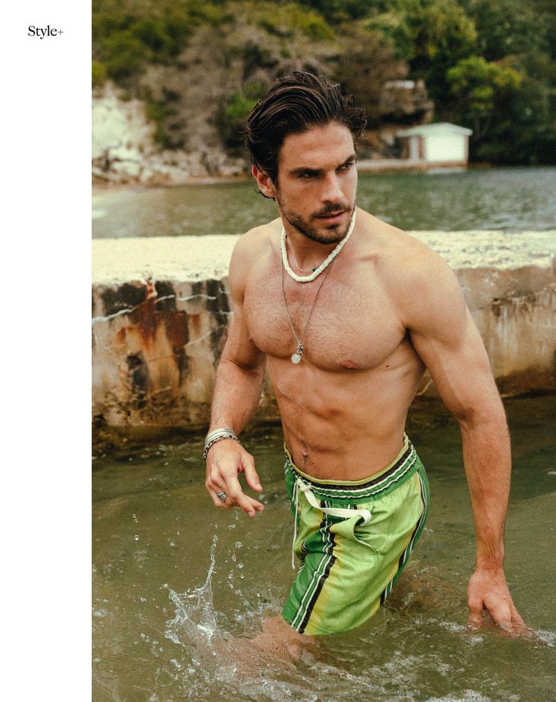 Handsome man with bare chest stands in shallow water wearing green swim trunks. The background features coastal trees and a building.