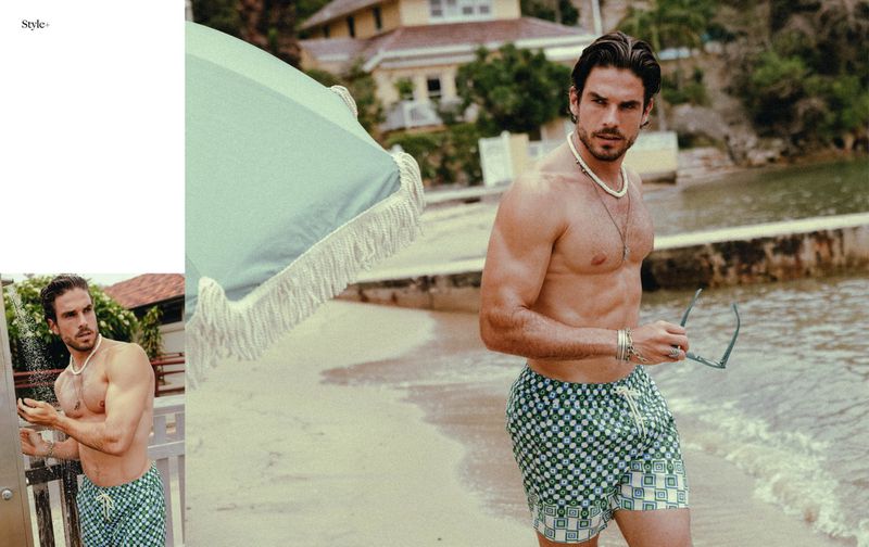 Shirtless man in patterned swim trunks standing on a sandy beach near an umbrella, sunglasses in hand, with a building in the background.