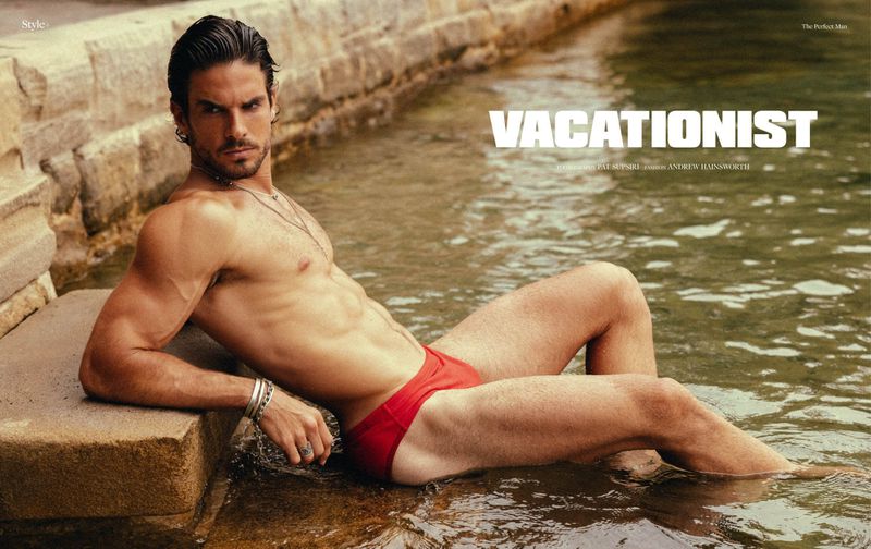 An adult male model poses while reclining on a stone surface in a body of water wearing red swimwear in an eye-level outdoor shot.