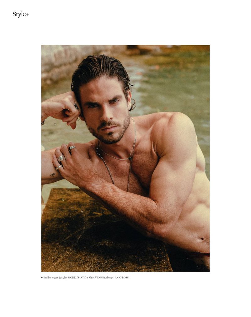 Shirtless male model poses with jewelry in a daylight studio shot, showcasing fashion and fitness in a neutral outdoor setting.
