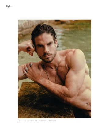 Shirtless male model poses with jewelry in a daylight studio shot, showcasing fashion and fitness in a neutral outdoor setting.