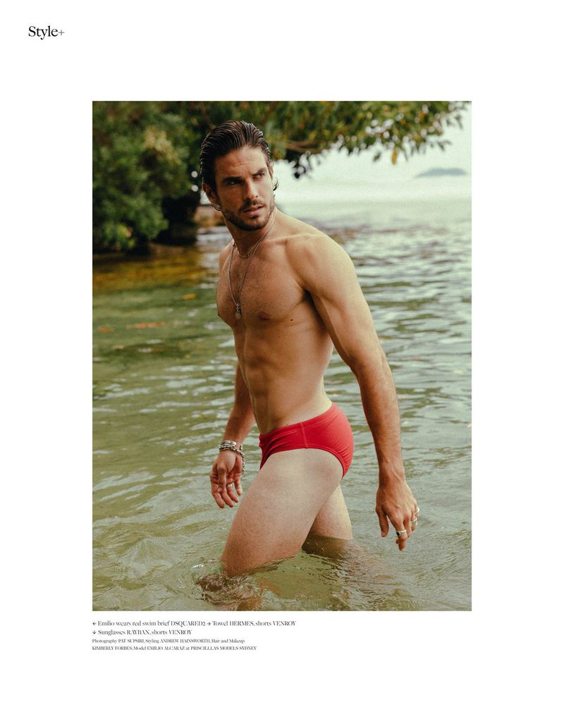 Athletic adult male in red swim trunks standing in ocean water with green trees in background on a sunny day in an outdoor setting.