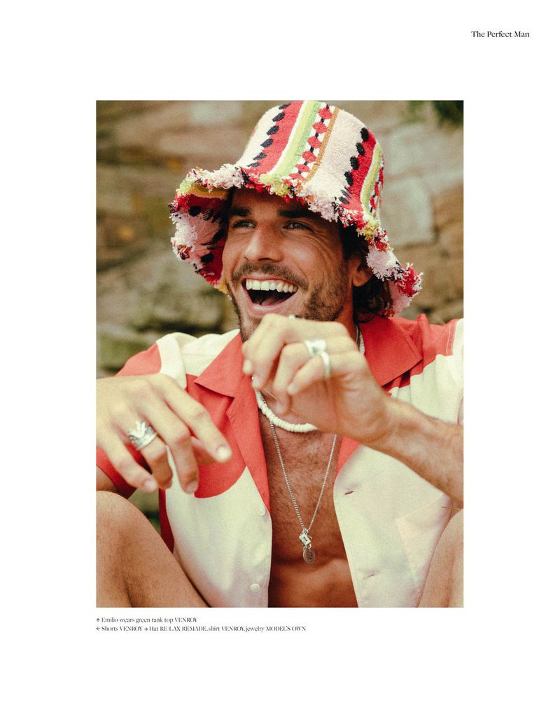 Portrait of a man with a colorful bucket hat laughing outdoors in a casual orange and white shirt, wearing rings and necklace.