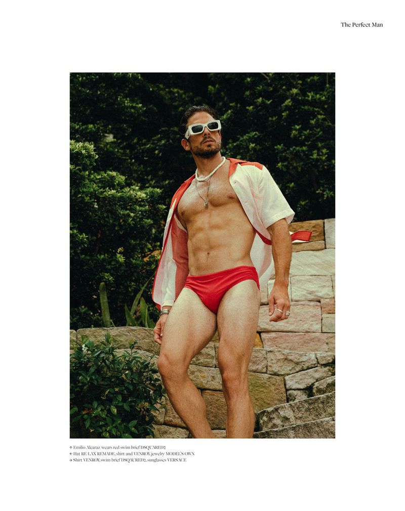 A man poses on stone steps outdoors, wearing a red swimsuit and unbuttoned shirt, complemented by sunglasses and a necklace.
