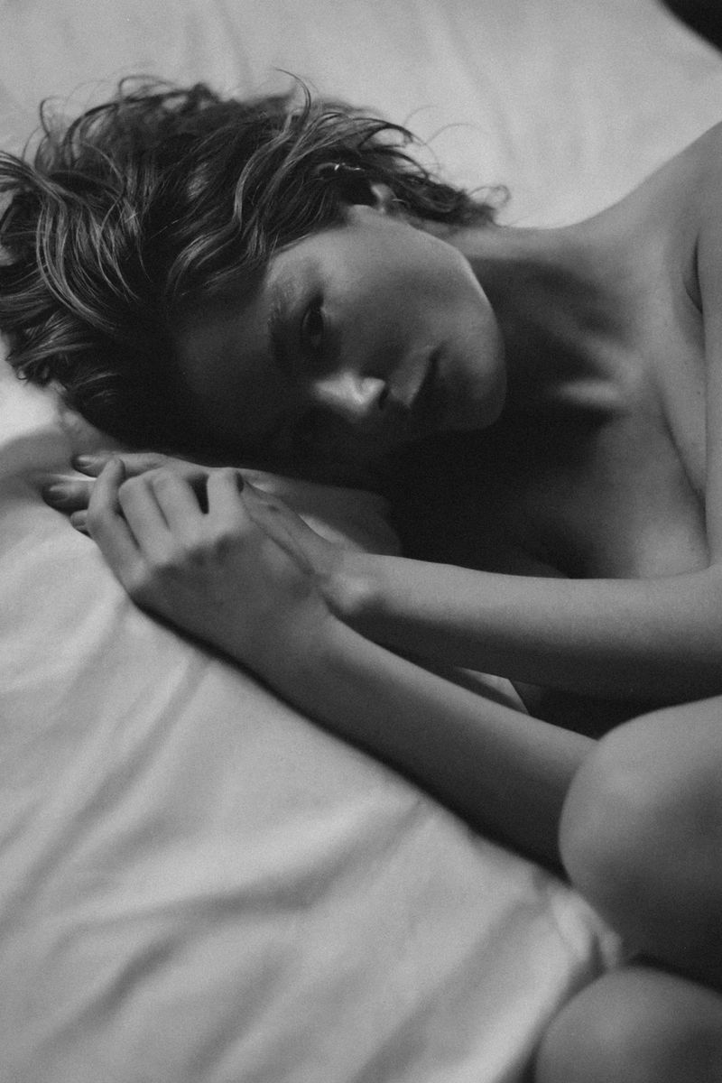 Black and white portrait of a young adult woman lying on a white bed with her head resting on the pillow and her hands folded.