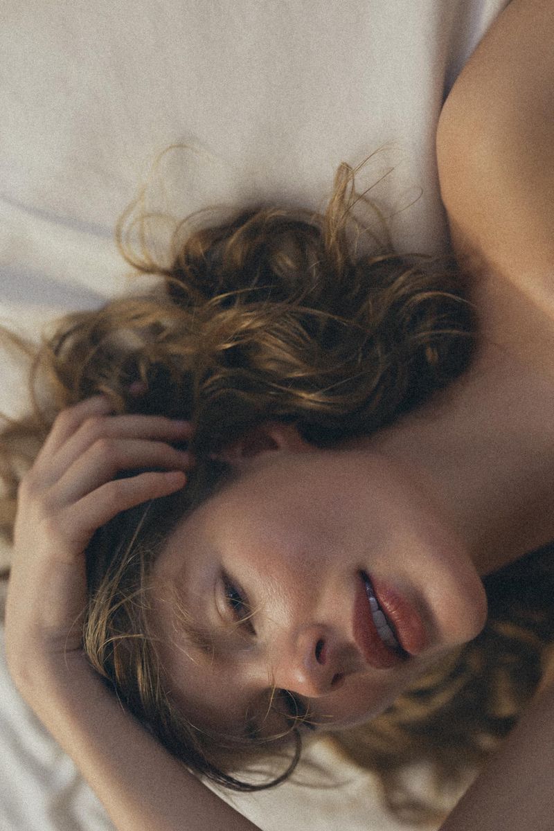Adult female lying on a white bed with her hand in her brown wavy hair, looking softly at the camera with a gentle expression.