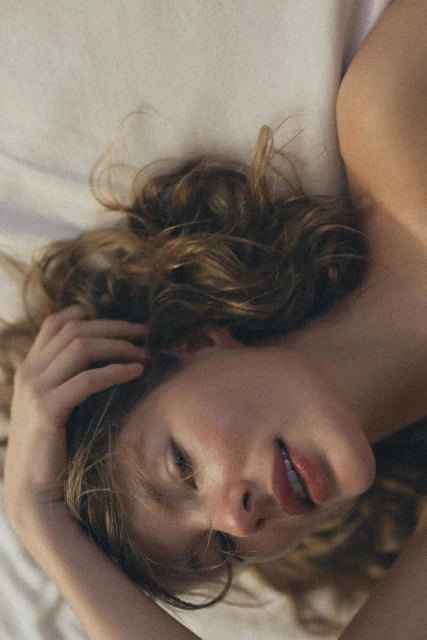 Adult female lying on a white bed with her hand in her brown wavy hair, looking softly at the camera with a gentle expression.