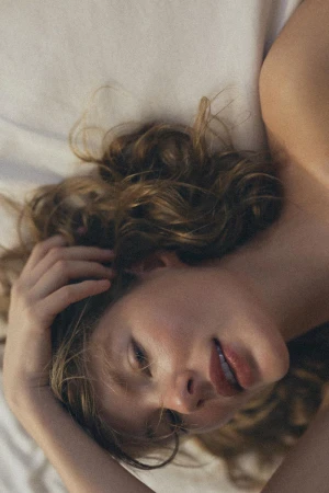 Adult female lying on a white bed with her hand in her brown wavy hair, looking softly at the camera with a gentle expression.
