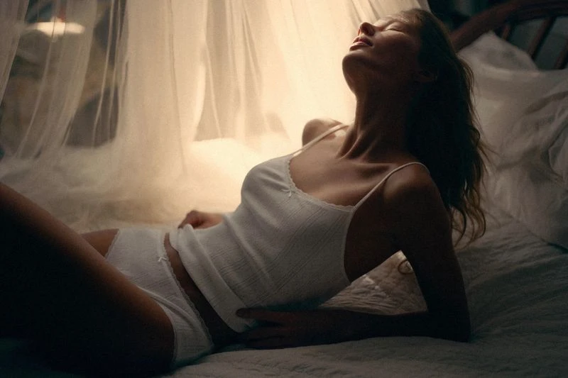 Woman reclines on a bed bathed in soft light, wearing a white camisole and underwear, under sheer white curtains in a gentle pose.