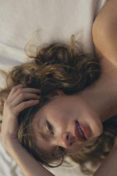 Close-up portrait of a woman with curly brown hair lying on white bedsheets, gently resting her hand in her hair.