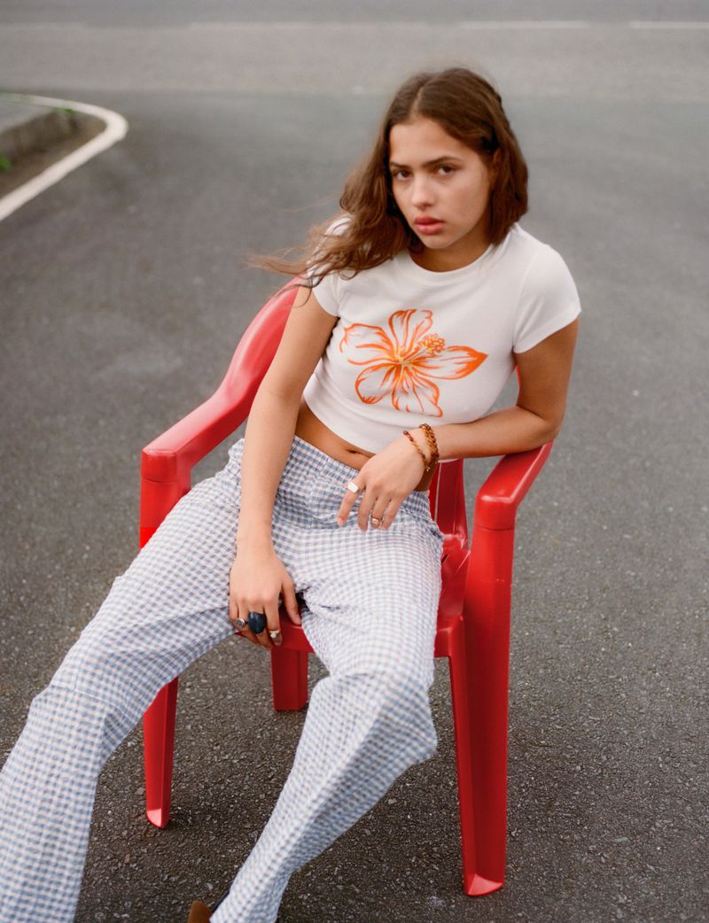 Young woman sits on a red plastic chair in the middle of a road, wearing a white t-shirt with an orange flower print and checkered pants.