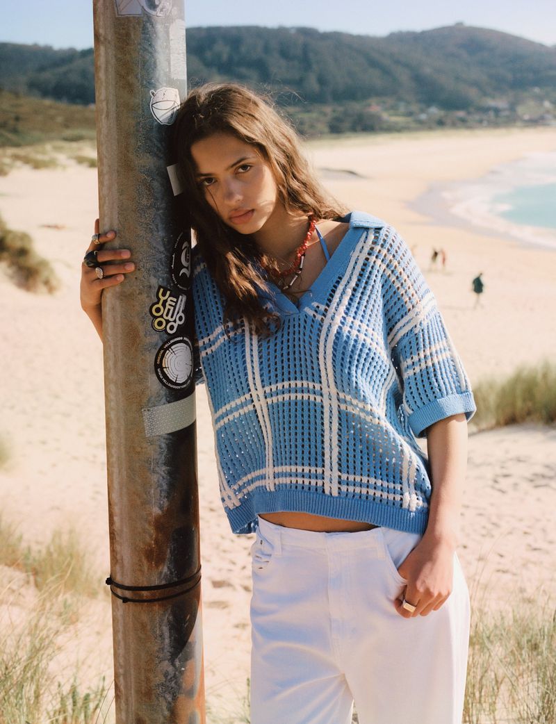 Woman in knit sweater leaning against pole at the beach, wearing white pants, jewelry, in a medium shot with a sunny day background.