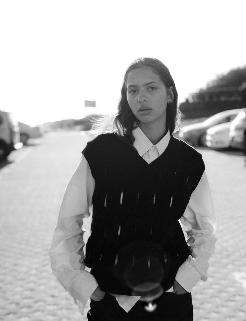 A young adult woman in a sweater vest stands outside in black and white with cars parked in the background on a sunny day.