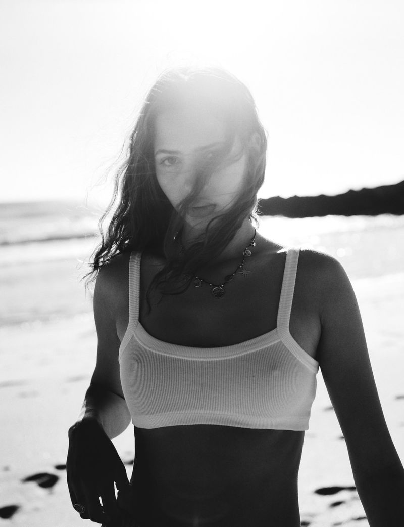 Black-and-white portrait of a young woman in a bralette on a beach, facing the camera with sunlight and ocean in the background.