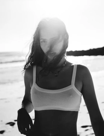 Black-and-white portrait of a young woman in a bralette on a beach, facing the camera with sunlight and ocean in the background.
