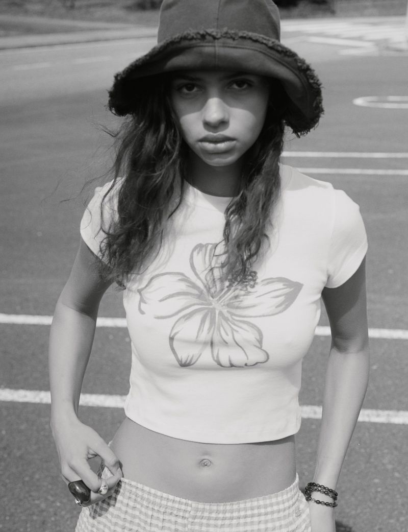 Black and white portrait of a young woman wearing a hat and a t-shirt with a flower graphic, standing outside on a paved surface.