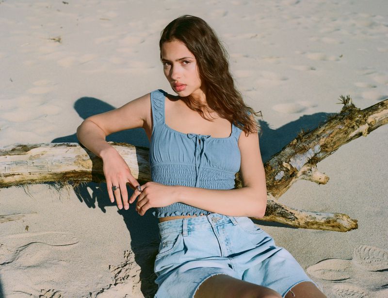 Young woman in a blue top and light blue denim shorts sitting on a log on a sandy beach in sunlight looking at the camera.