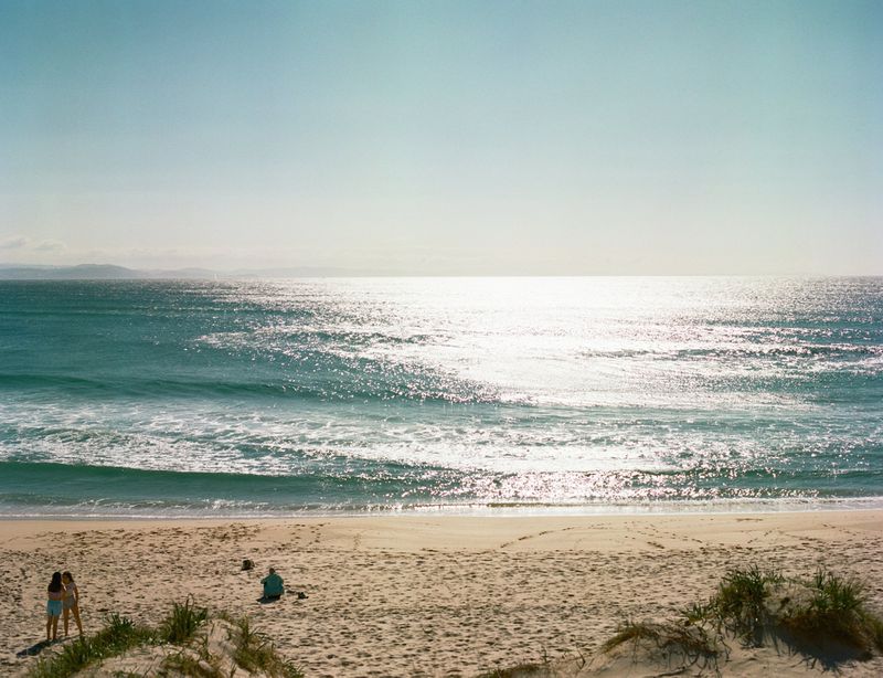Ocean view of turquoise waves meeting sandy beach with two women standing and another person sitting at shore, under sunny sky.