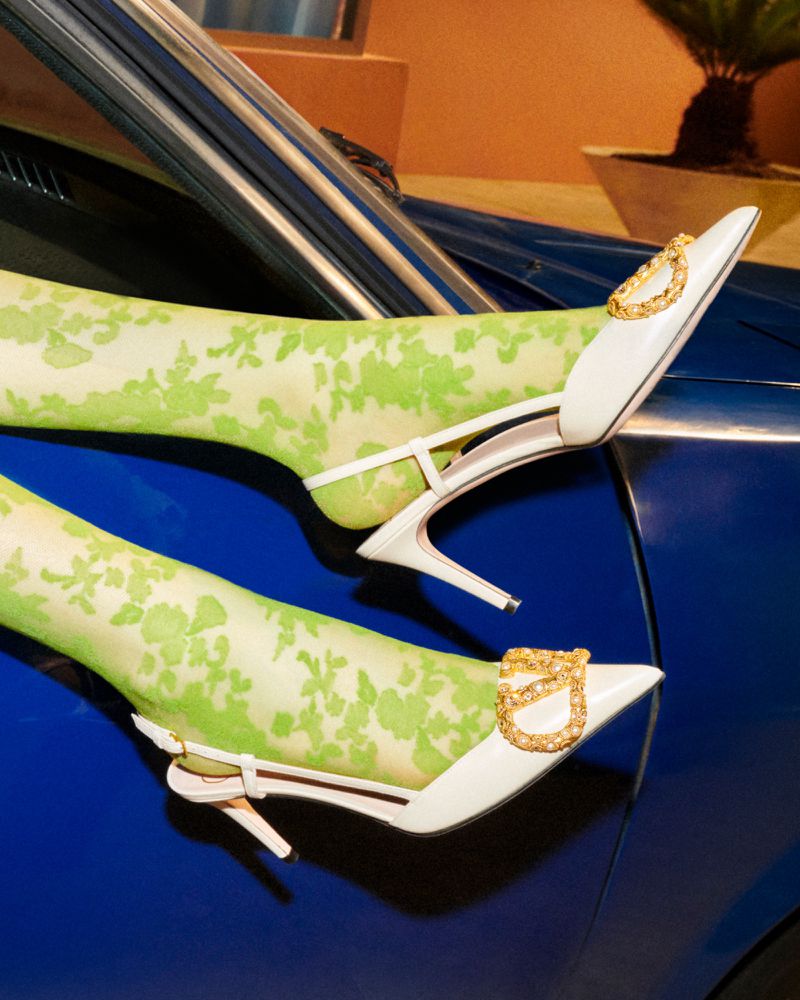 Elevated view of white slingback heels with jeweled buckle, worn with patterned hosiery, resting on the glossy hood of a blue car.