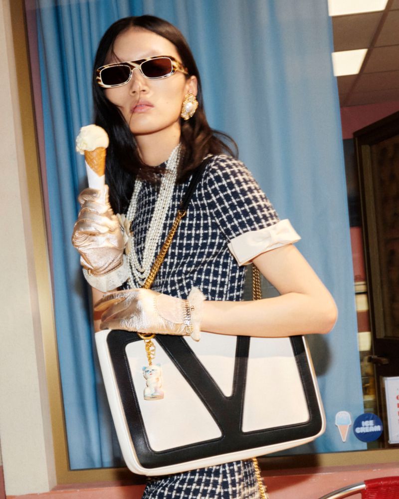 An Asian woman poses with an ice cream cone, wearing sunglasses, a dress, jewelry, gloves, and holding a purse.