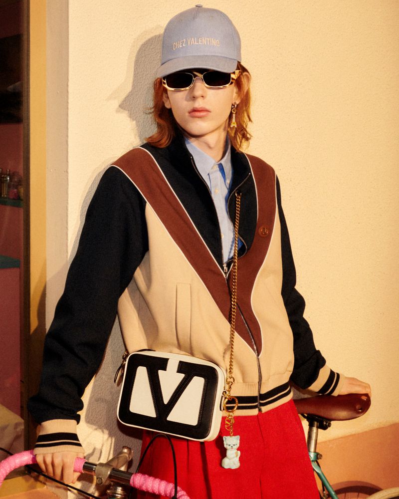 An adult male poses with a bicycle in an indoor studio with a light blue baseball cap, sunglasses, and a shoulder bag with a 'V' on it.