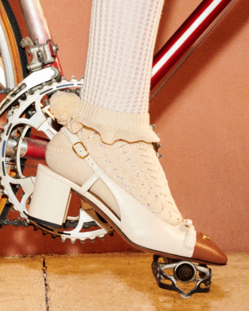 Close-up of a foot wearing a two-tone heel and a ruffled sock, resting on a bicycle pedal, with the bike frame in the background.