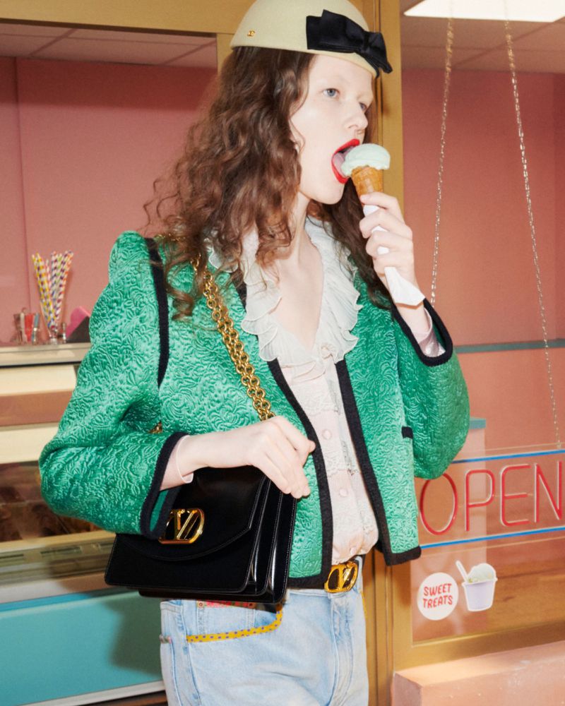 Fashion shot of a model eating ice cream, wearing a green jacket, hat, and jeans, accessorized with a black handbag and gold jewelry.