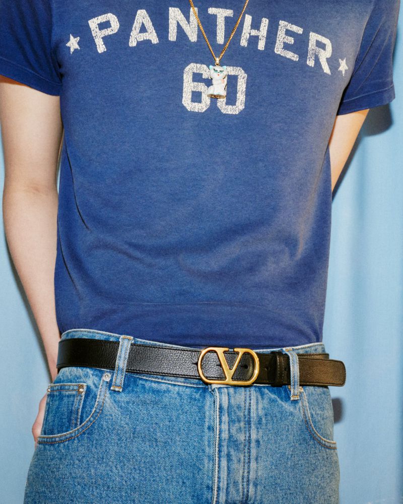 Close-up of a person wearing a blue t-shirt, jeans, and a Valentino belt with a gold buckle against a light blue background.