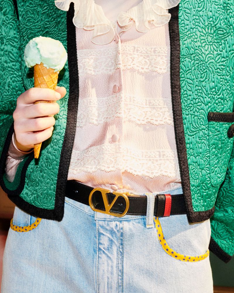 Close-up of a person holding an ice cream cone, wearing a green jacket and jeans with a Valentino belt, casual style, fashion shoot.