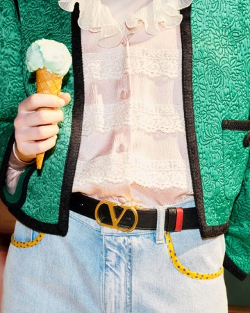 Close-up of a person holding an ice cream cone, wearing a green jacket and jeans with a Valentino belt, casual style, fashion shoot.