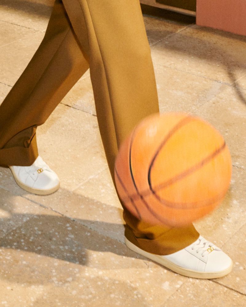 Person in tan trousers and white sneakers bouncing a basketball on a concrete surface in neutral light. Focus on the shoes and ball.