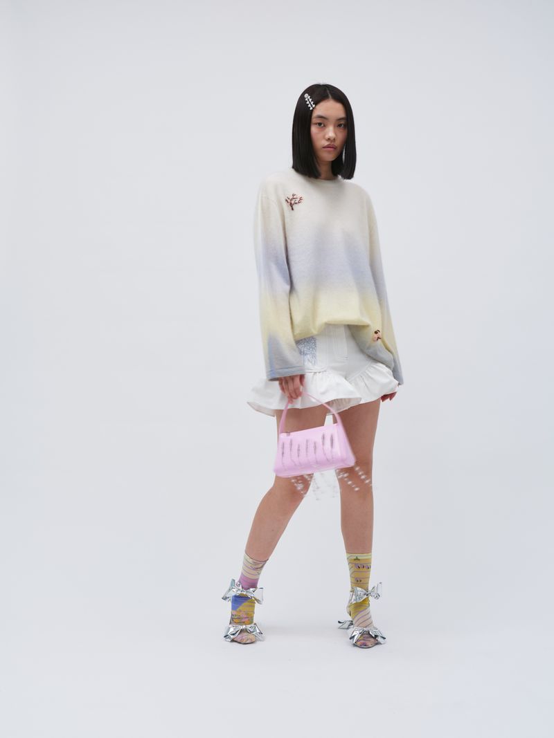Woman poses in pastel sweater and white skirt with pink purse in studio lighting. Fashion shot with soft colors and unique accessories.