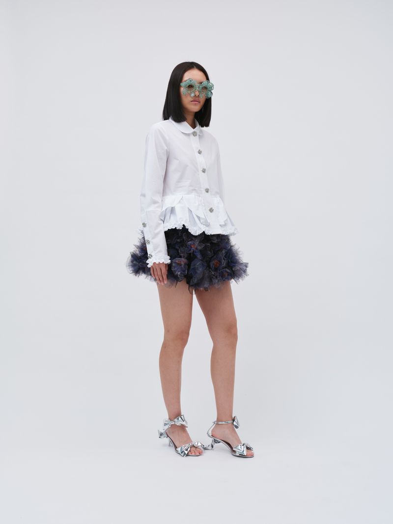 Young woman in fashionable outfit with sunglasses, a white long-sleeved blouse, a frilly dark miniskirt, and glittery heels on a white background.
