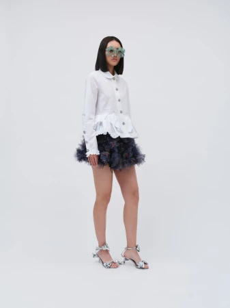 Young woman in fashionable outfit with sunglasses, a white long-sleeved blouse, a frilly dark miniskirt, and glittery heels on a white background.